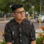 A man with glasses sits in a park wearing a patterned shirt and using earbuds.